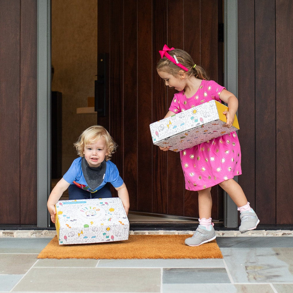 Two children with boxes on a stone patio in front of a wooden door.