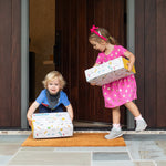 Two children with boxes on a stone patio in front of a wooden door.