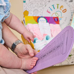 A baby's foot is being measured on a purple plastic measuring tool held by an adult, with a colorful box of baby socks in the background.