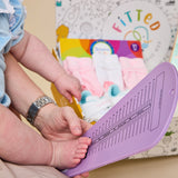 A baby's foot is being measured on a purple plastic measuring tool held by an adult, with a colorful box of baby socks in the background.