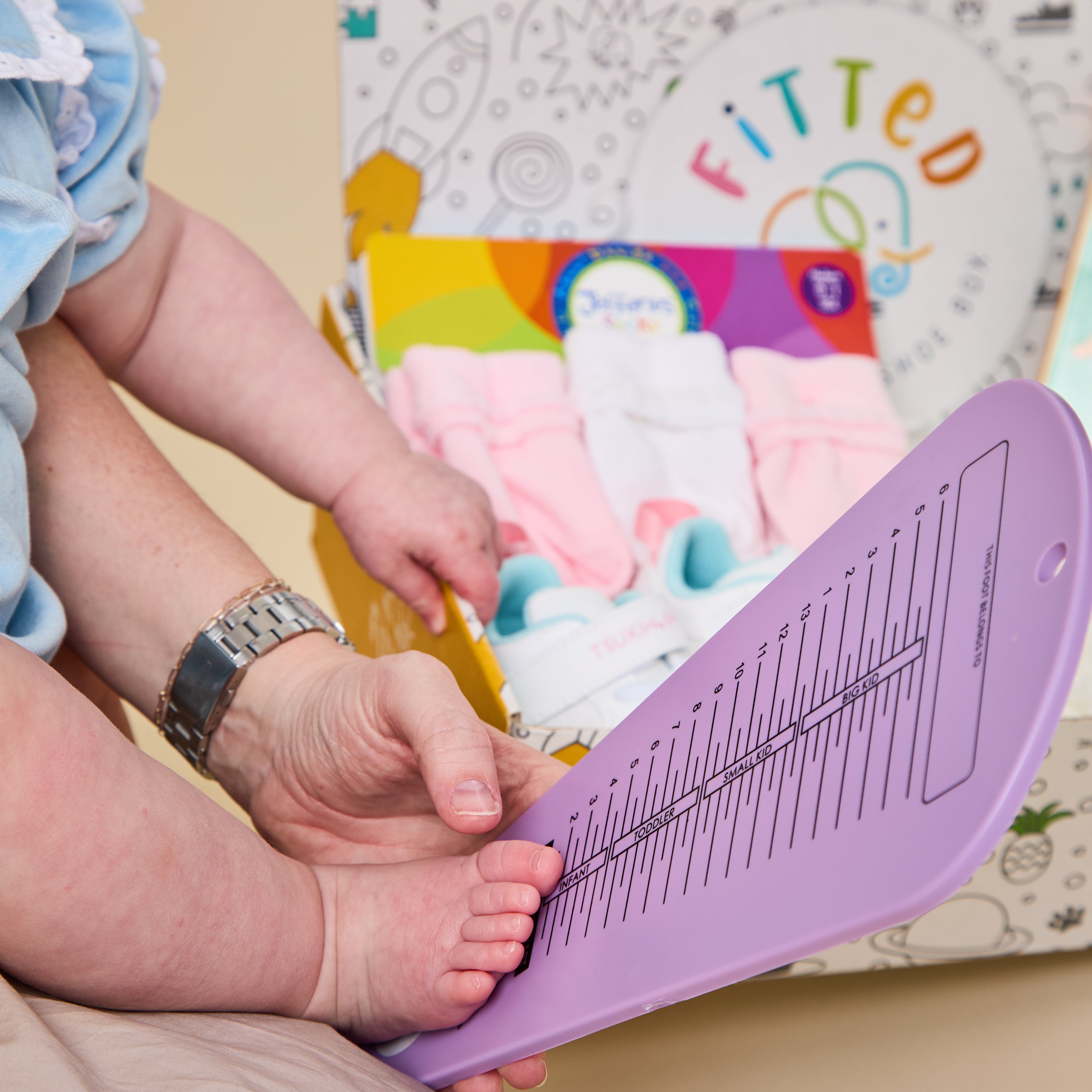 A baby's foot is being measured on a purple plastic measuring tool held by an adult, with a colorful box of baby socks in the background.