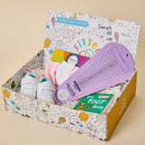 A baby foot gift box in a decorative cardboard box, featuring a purple measuring tool, white and blue baby shoes, pink socks, a colorful book, and a green 'The Foot Book' on a beige background.