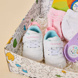 A baby gift set in a patterned box containing a pair of white TSUKIHASHI baby sneakers with pastel pink and light blue accents, pink and white socks, and a purple FIT FLOW yoga mat on a beige background.