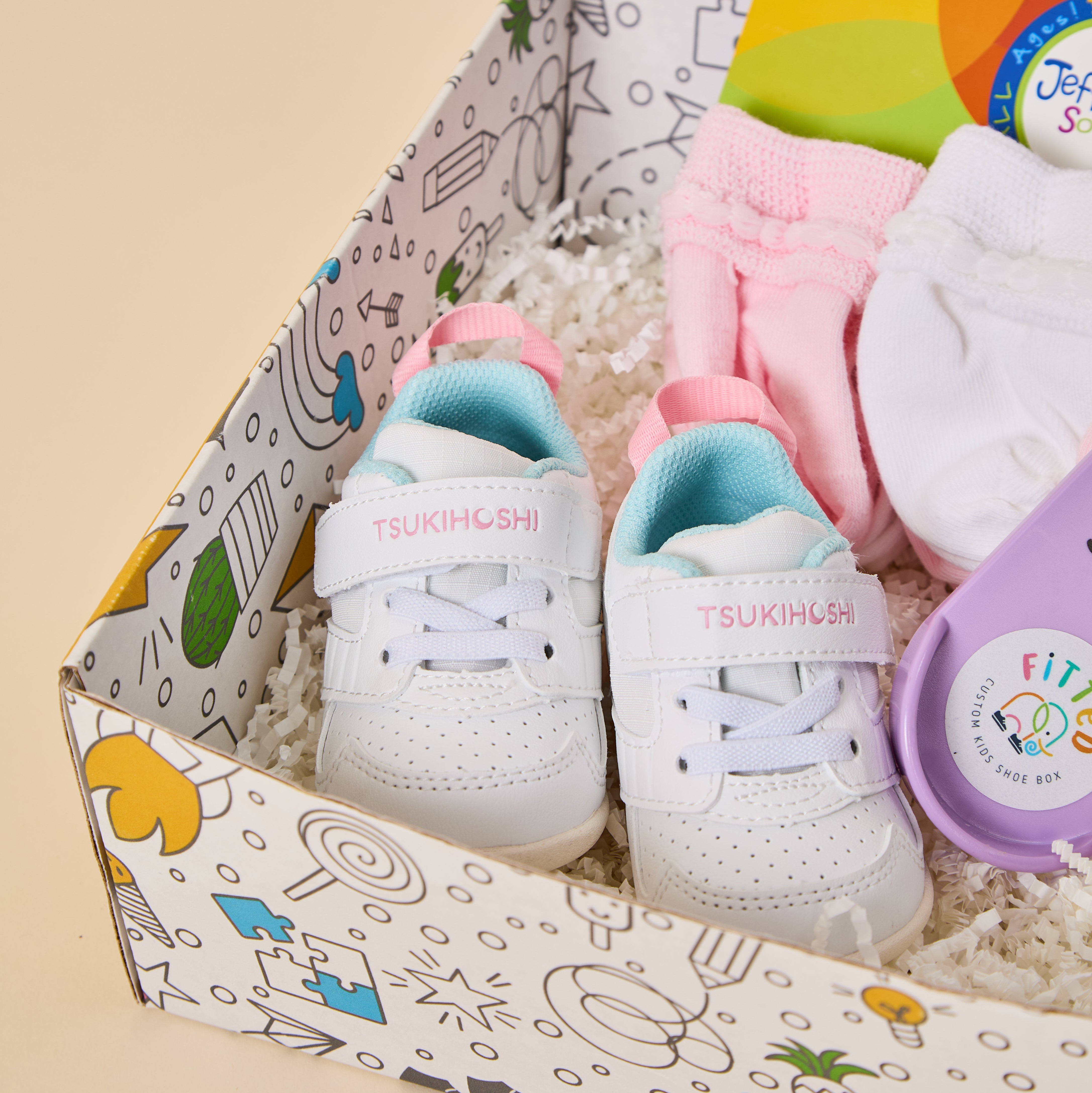A baby gift set in a patterned box containing a pair of white TSUKIHASHI baby sneakers with pastel pink and light blue accents, pink and white socks, and a purple FIT FLOW yoga mat on a beige background.