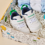 A pair of white baby sneakers with green and navy accents, displayed inside a decorative gift box with shredded paper filler and folded socks.