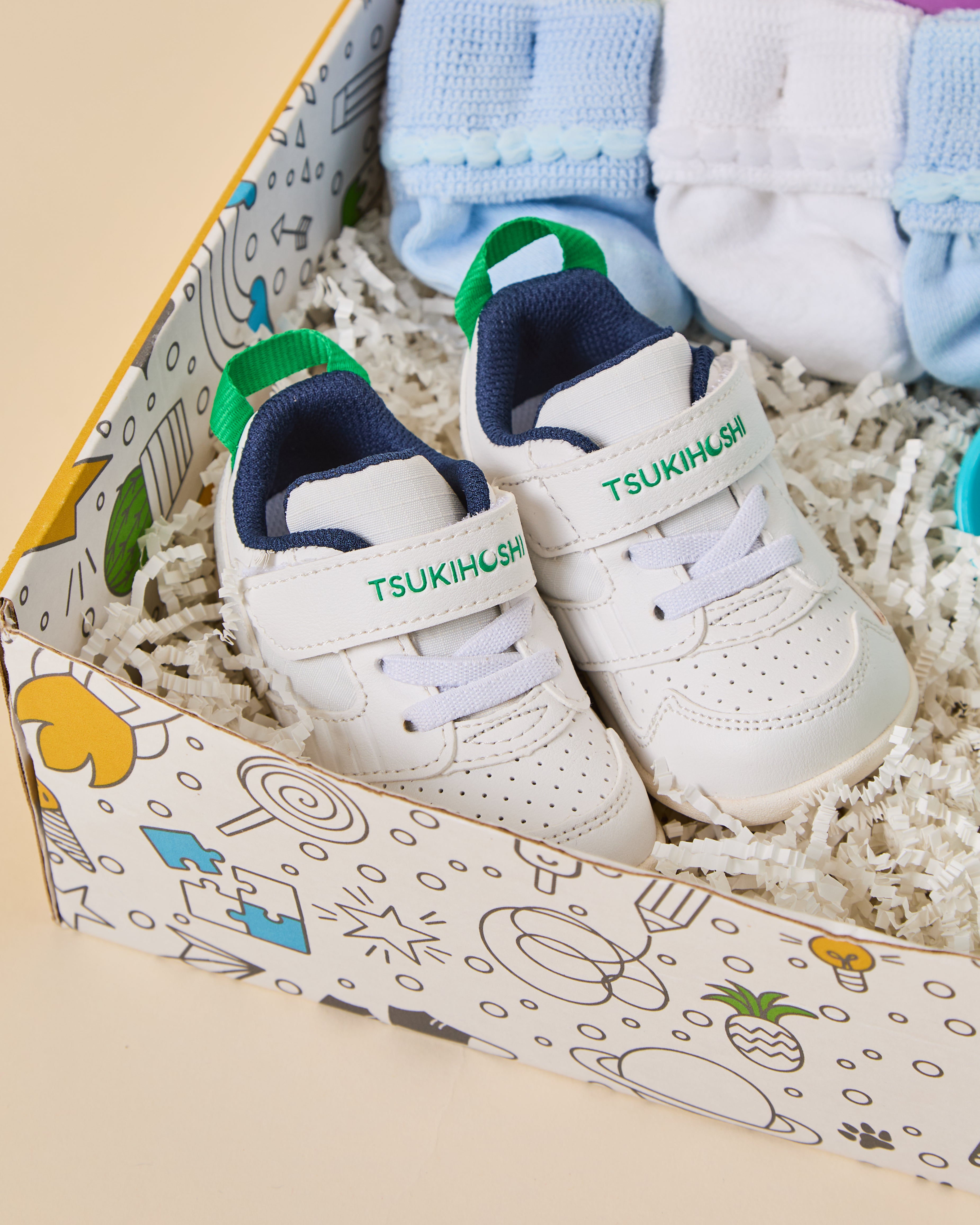 A pair of white baby sneakers with green and navy accents, displayed inside a decorative gift box with shredded paper filler and folded socks.