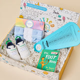 A curated gift box for children's feet and toes, filled with white sneakers, pastel socks, a blue foot measuring tool, a Dr. Seuss book, and folded socks, set against a solid beige background.